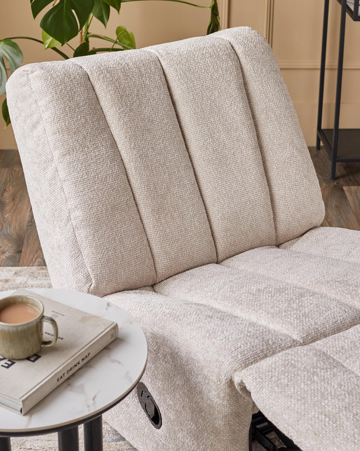 A textured, off-white recliner chair sits next to a round, marble-topped side table with a book and cup on it.