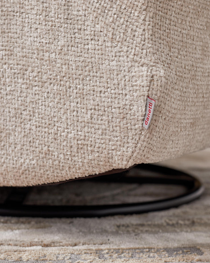 Close-up of a textured, beige upholstered chair with a 'danetti' tag, and a glimpse of the swivel base and patterned rug beneath.