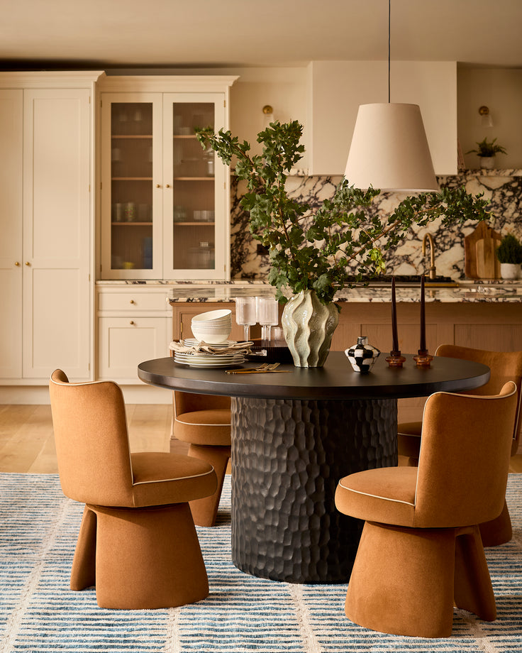 Knox black round dining table with textured pedestal base, paired with cognac leather chairs in a cream kitchen setting.