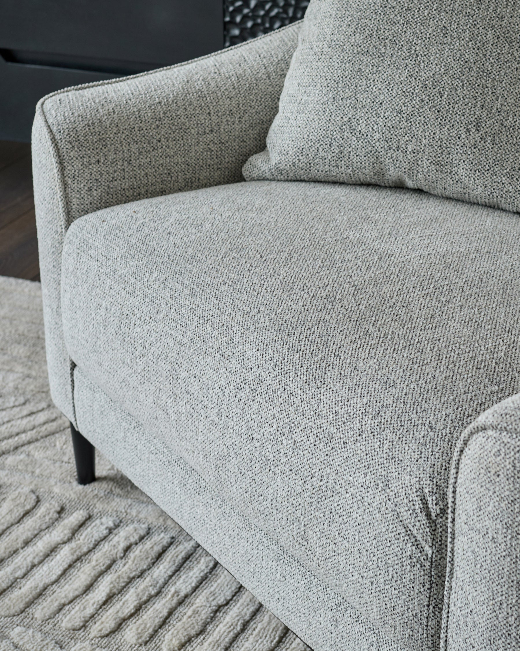 Grey fabric armchair with black wooden legs on striped grey rug, angled side view showing curved arm and seat detail.