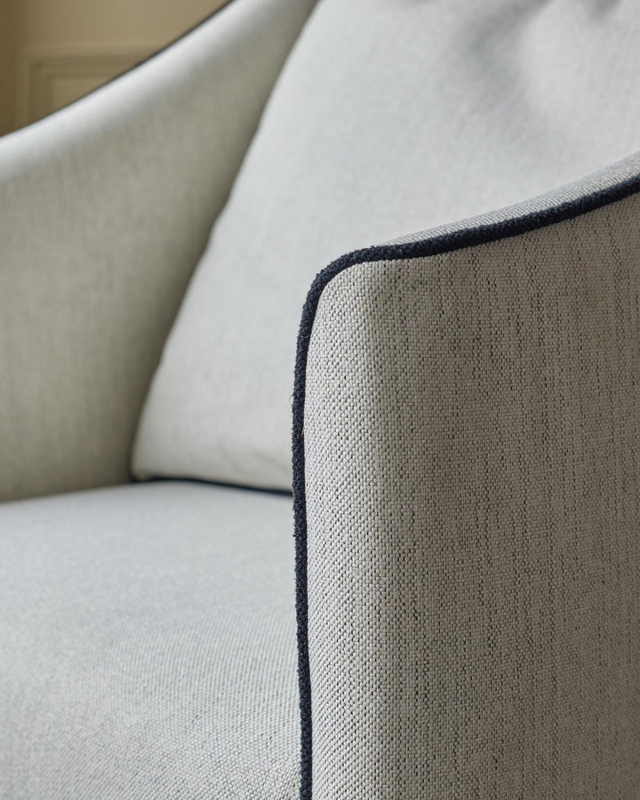 Close-up of a modern armchair in light grey fabric with dark grey piping around the armrest and seat cushion.