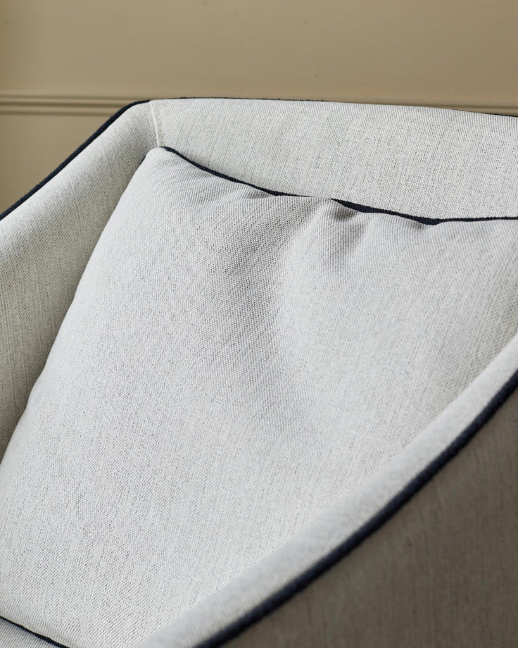 A close-up of a light grey armchair with dark blue piping around the edges and a matching cushion.