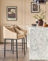 Honey fabric bar stool with black metal legs and brass footrest, shown at marble kitchen island in elegant interior setting.