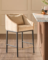 Julep bar stool in honey fabric with black metal legs and brass footrest, positioned at a marble-top counter.