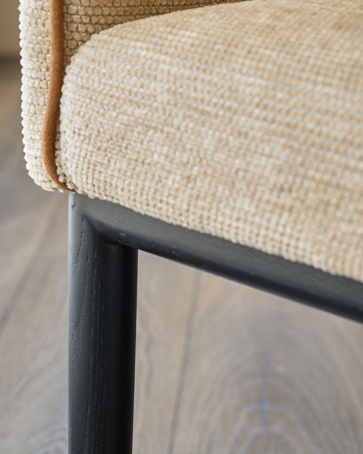 Julep Dining Chair in honey fabric, close-up of seat edge showing woven texture and wooden leather handle detail on black metal frame.