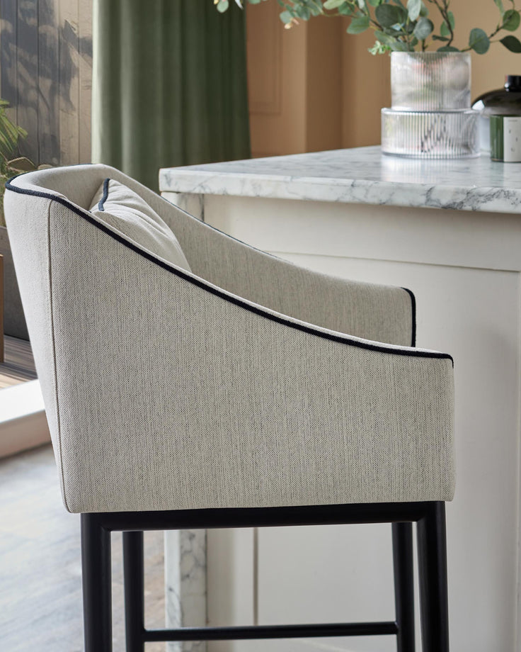 A modern bar stool with a light coloured cushioned seat and a black metal frame, positioned next to a marble-topped counter.