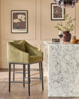 Olive-green upholstered bar stool with black wooden legs and brass footrests, shown in a styled interior setting.