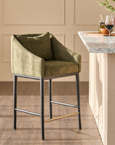 Olive green upholstered bar stool with dark metal legs and brass footrest, positioned at a marble-top counter.