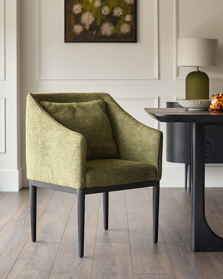 Olive green upholstered dining chair with black wooden legs, shown in a styled interior setting next to a dark side table.