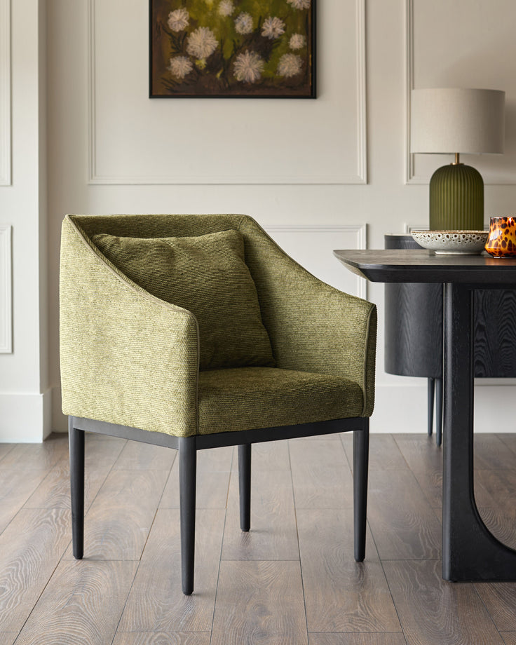 Olive green upholstered dining chair with black wooden legs, shown in a styled interior setting next to a dark side table.