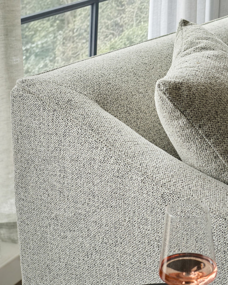 Grey woven fabric three-seater sofa with rolled arms, shown from a side angle near a window.
