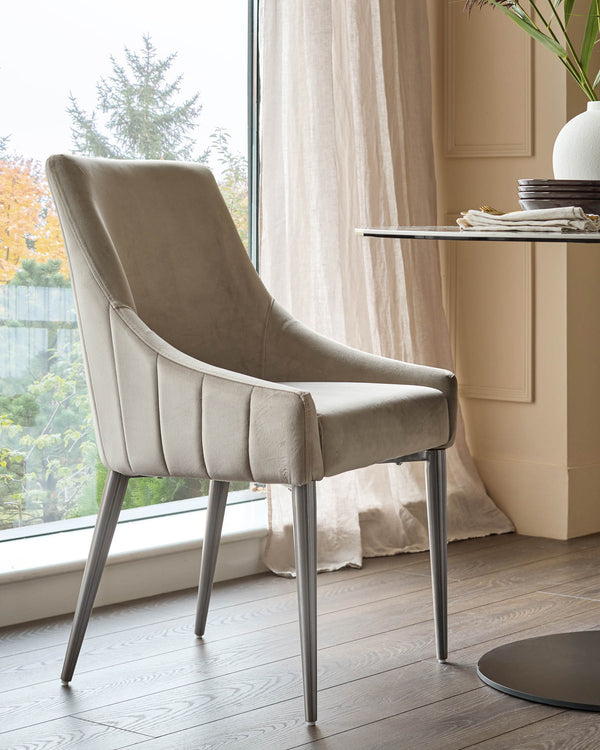 Champagne velvet dining chair with curved back, pleated sides, and stainless steel legs, positioned by a window.