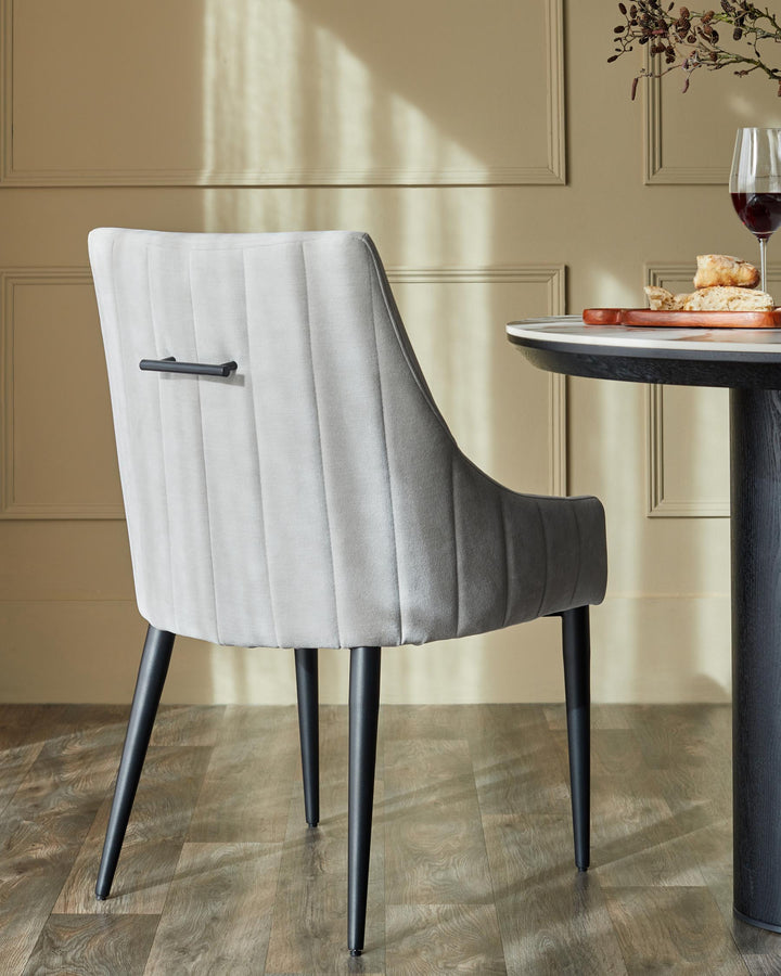 A grey upholstered dining chair with dark legs sits beside a modern round table with marble effect top and wooden base.
