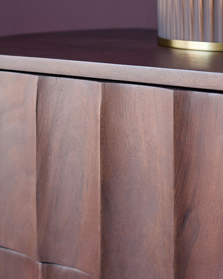 Dark mango wood bedside table with vertical panel detailing and brass hardware accent, angled side view.