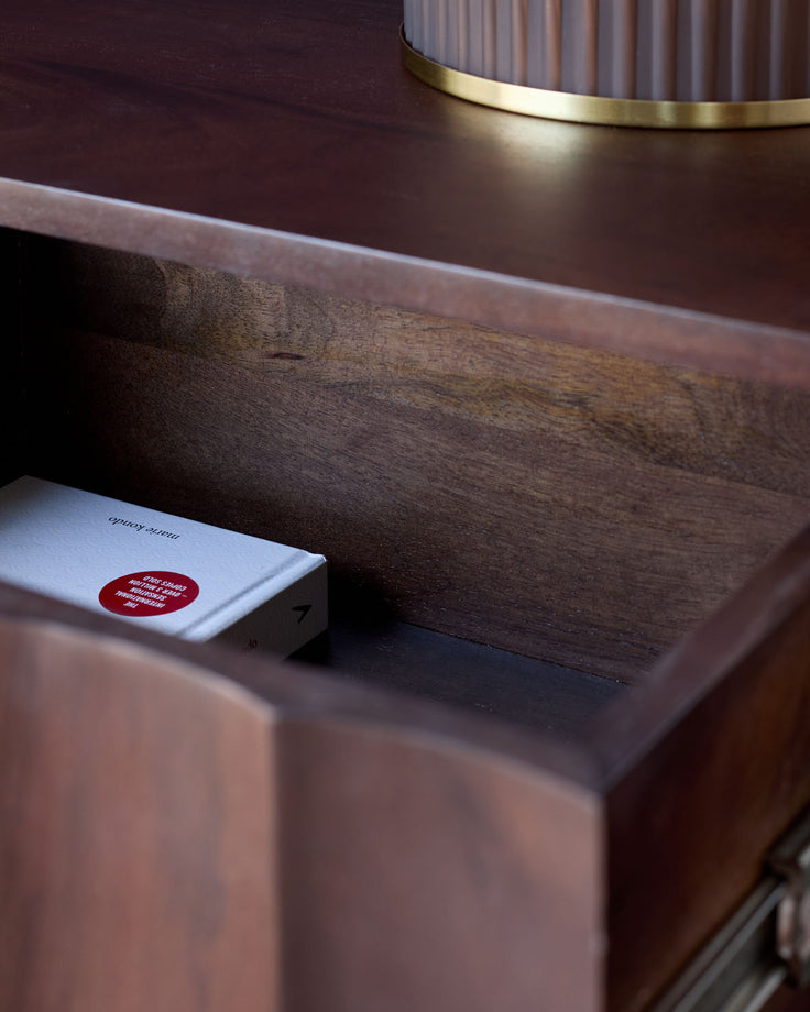 Dark mango wood bedside table with brass trim, featuring a drawer with a red circular label, angled side view.