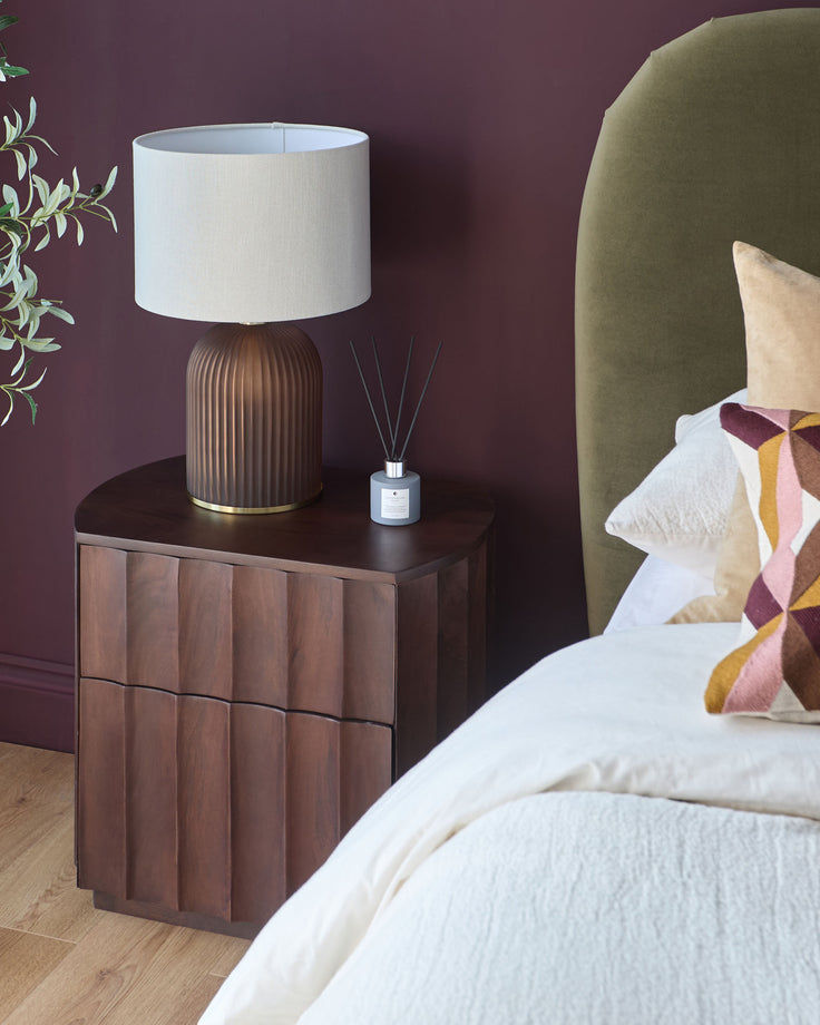 Juniper bedside table in dark mango wood with vertical paneled drawers, displayed in a bedroom setting next to a bed.