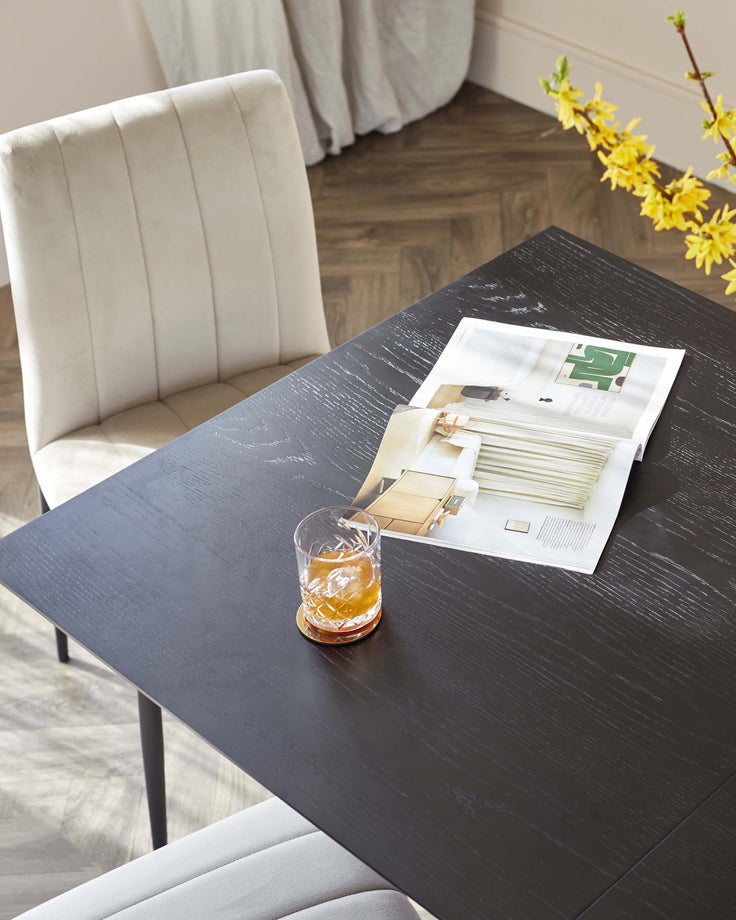 A dark wood dining table with modern beige chairs, a glass of whisky, and a magazine, creating a stylish and inviting dining space.