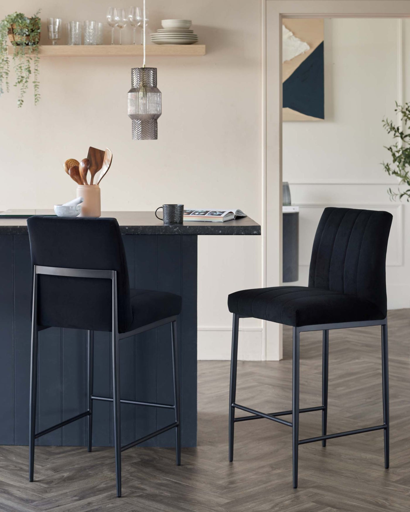 Two elegant bar stools with black upholstered seats and vertical channel stitching. The stools feature a high-back design and rest on slender metal legs with footrests. They are positioned against a dark modern kitchen island.