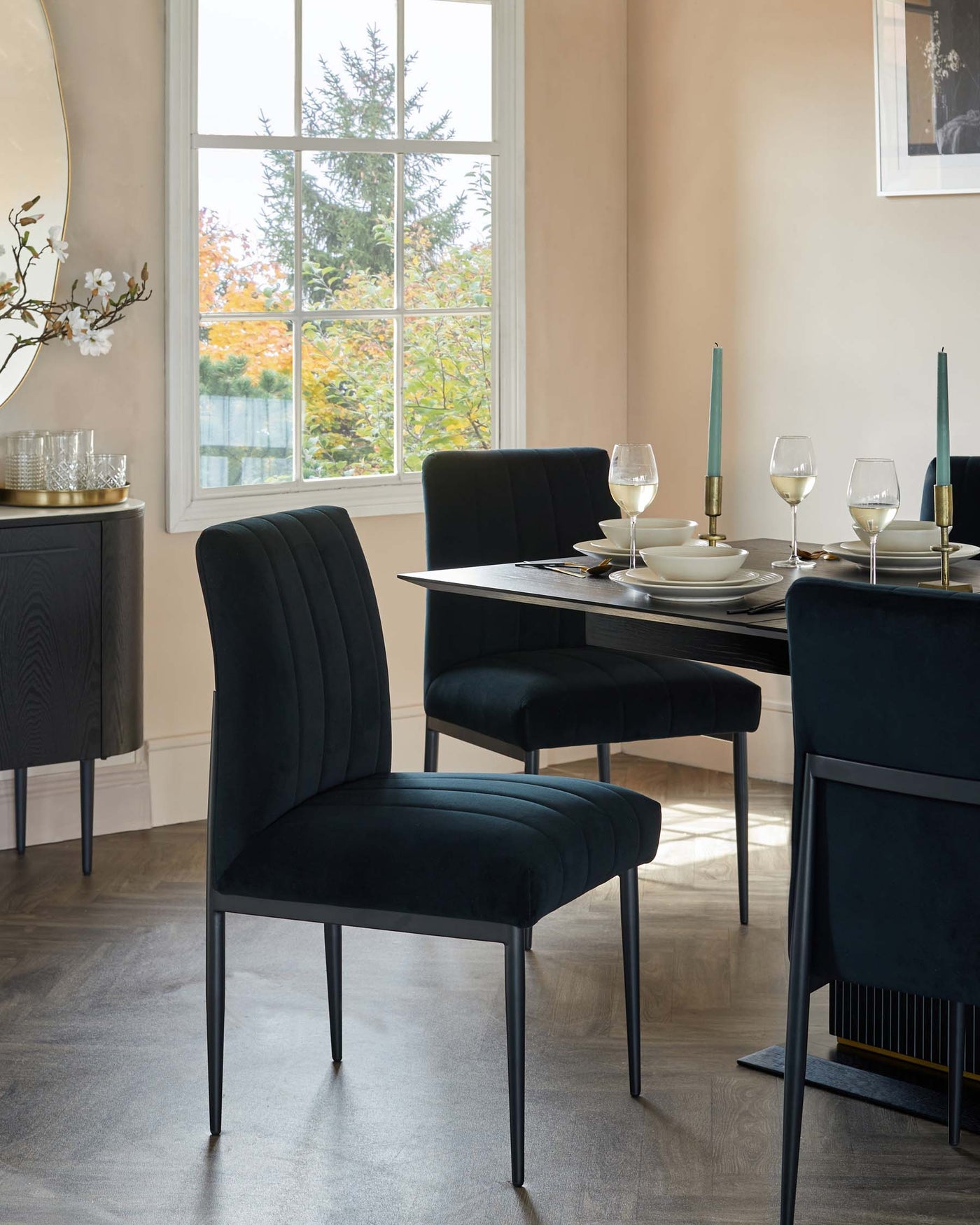 Elegant dining room furniture set featuring a modern rectangular black table and four plush velvet-upholstered chairs with vertical channel stitching and slim black metal legs. A coordinating dark wood sideboard with subtle geometric detailing and metal accents completes the sophisticated ensemble.