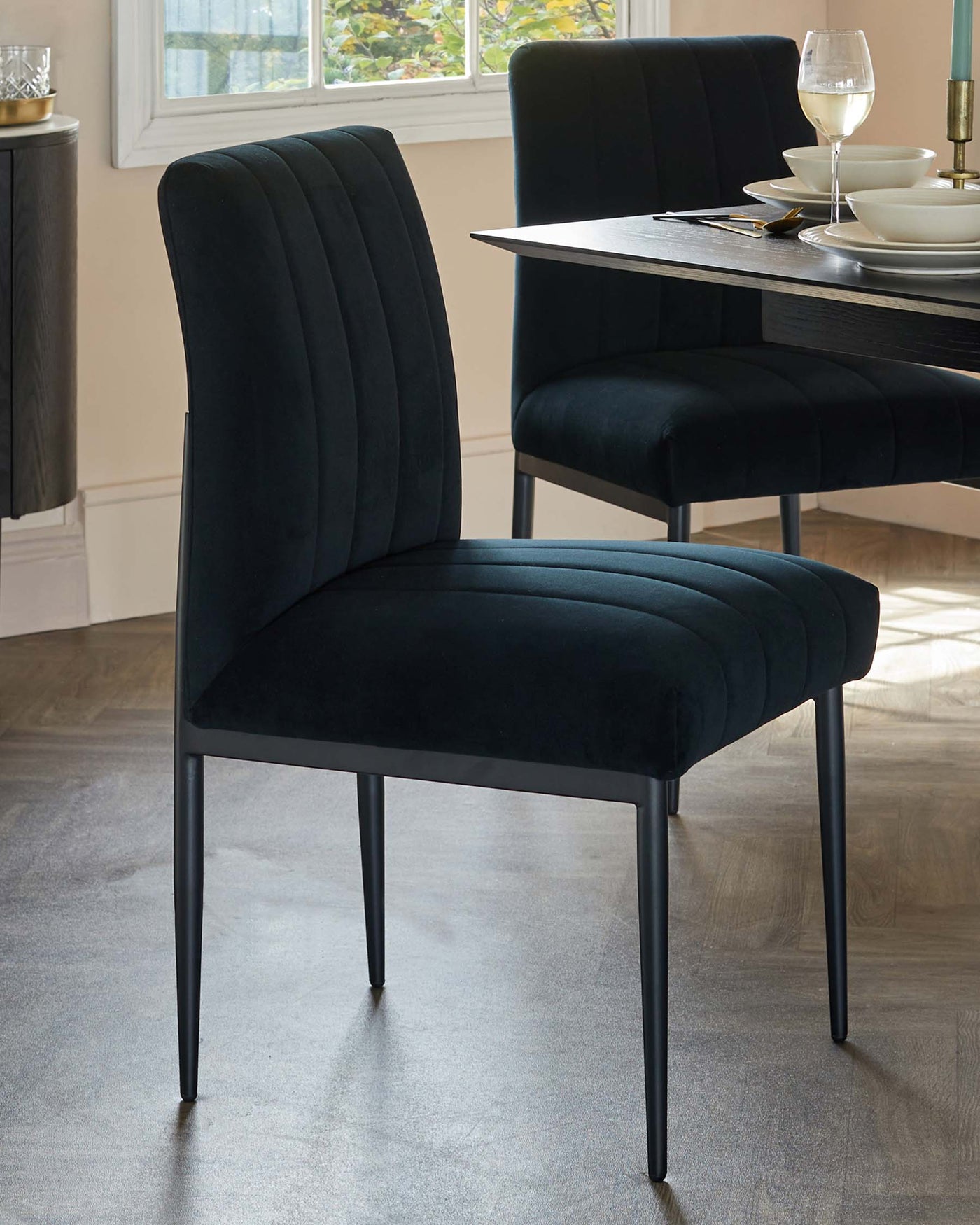 Elegant dining setting featuring modern velvet upholstered chairs with vertical channel tufting on the backrest and sleek black metal legs, complemented by a contemporary black dining table with a minimalist design, set against a room with natural light and a neutral colour palette.