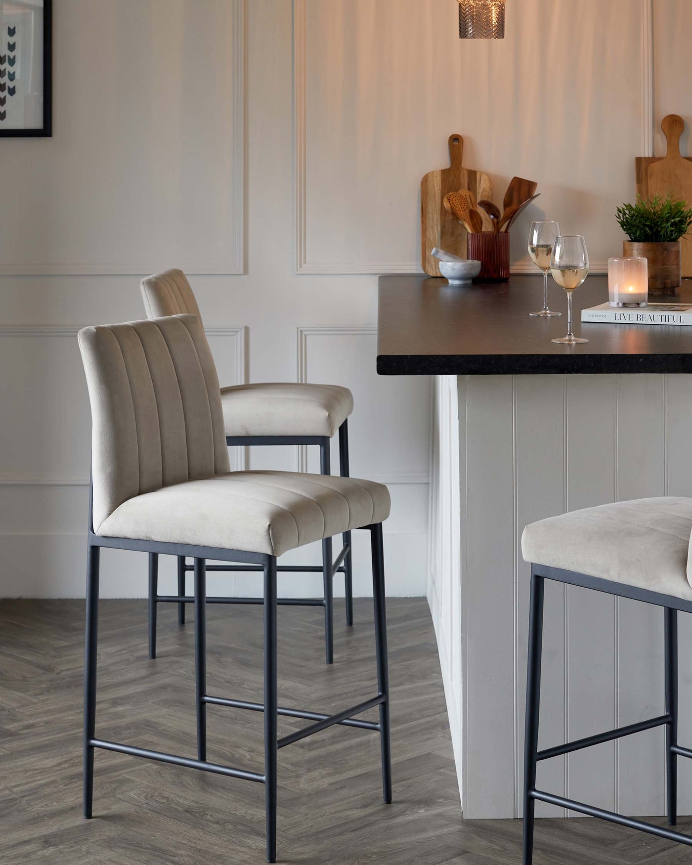 Elegant modern bar stools with beige velvet upholstery and channel tufting on the backrest, seated on a sleek black metal frame with a footrest, positioned adjacent to a kitchen island with a dark countertop.