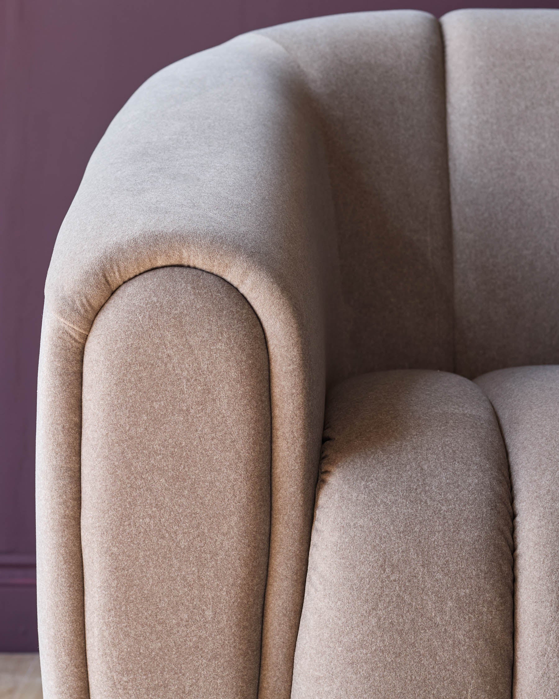 Close-up of a beige upholstered sofa with curved armrests against a purple wall, showcasing modern design and comfort.