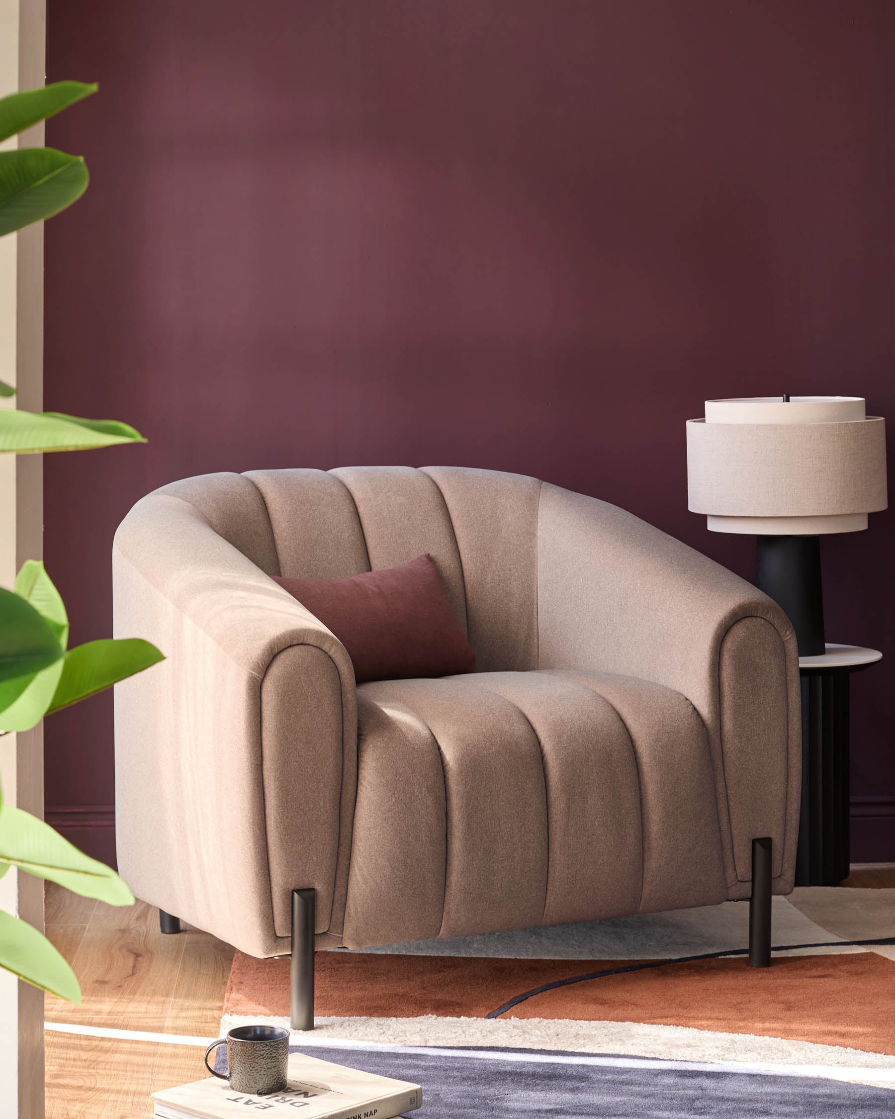 A contemporary neutral armchair with a brown cushion sits on a colourful rug, next to a lamp and a houseplant.