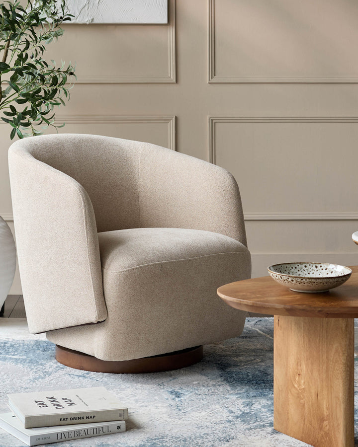 Beige swivel chair next to a small round wooden coffee table, books and a plant are visible on a light blue rug.