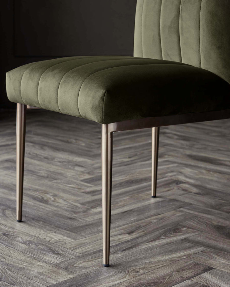 A modern velvet chair in olive green, with a vertically-stitched back and gold-tone legs, stands on a herringbone-patterned floor.