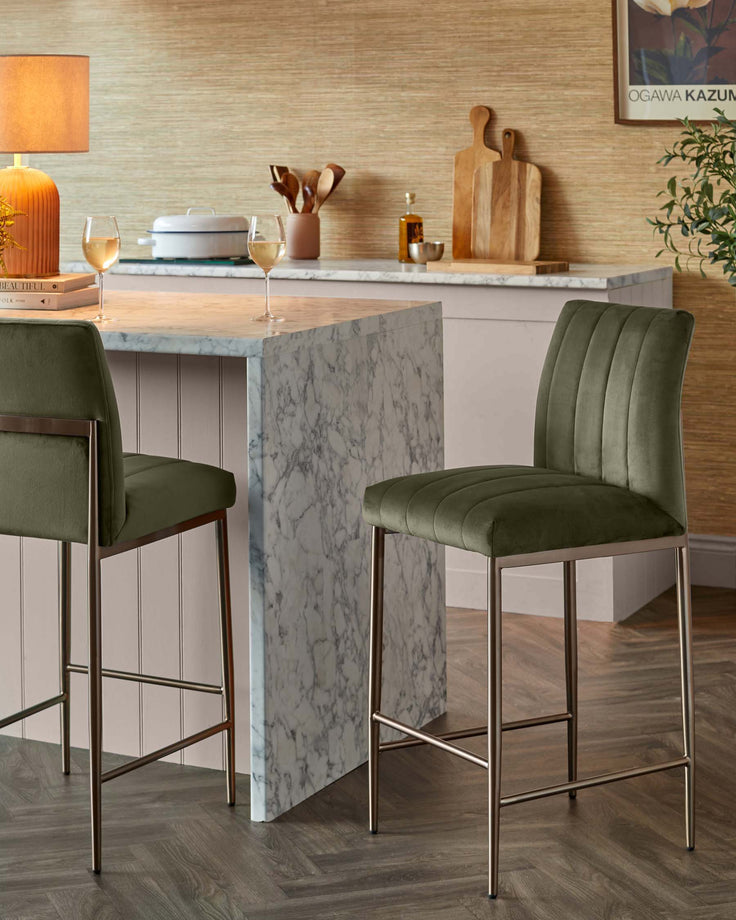 Two olive green velvet bar stools with sleek metal frames sit in a stylish kitchen with a marble island.