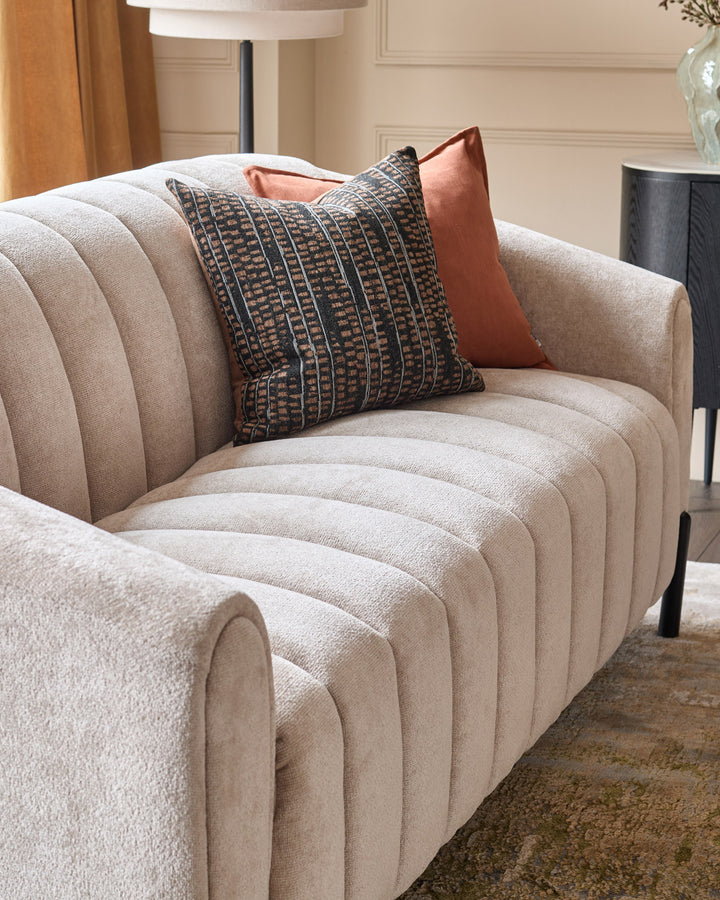 Kendra 3-seater sofa in natural fabric, featuring curved channeled design with decorative pillows in a modern living room setting.