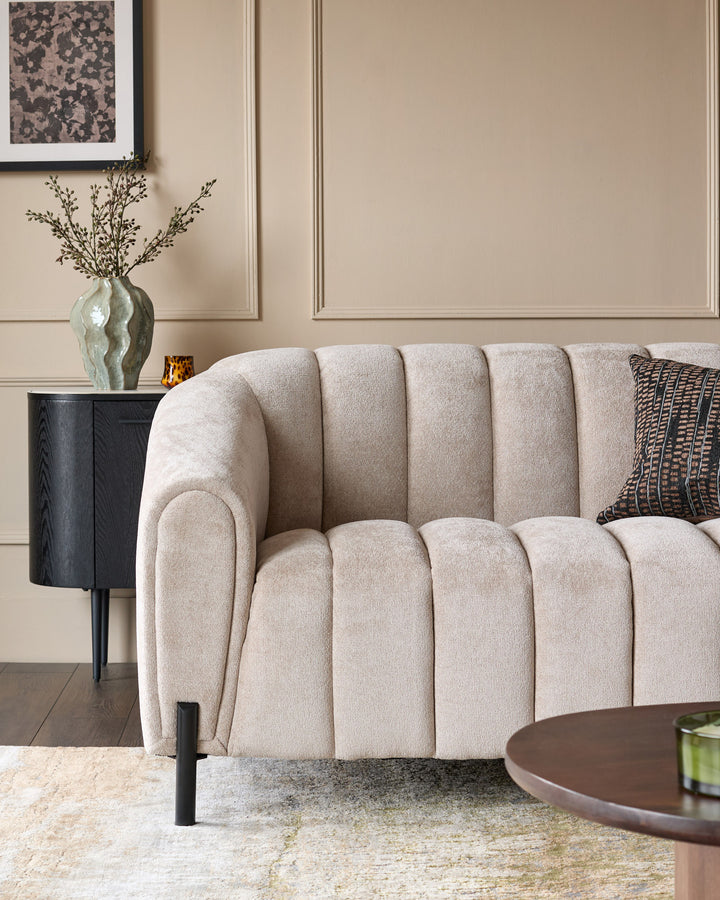 Kendra 3-seater sofa in natural beige fabric with vertical channel quilting, black legs, displayed in a styled living room setting.