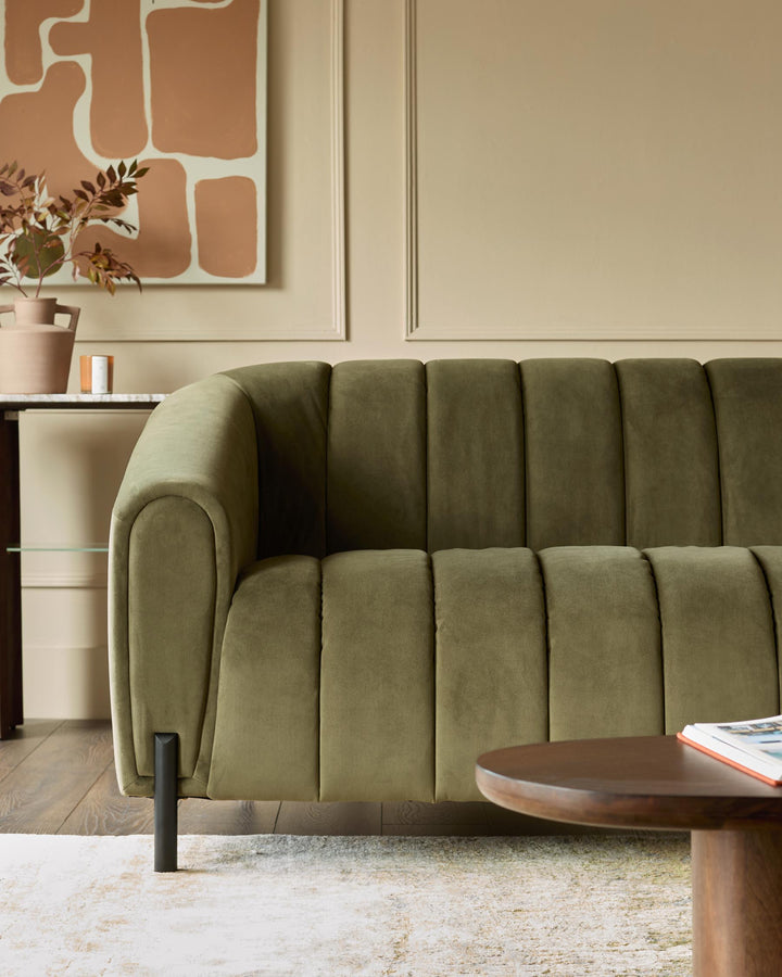 A stylish, olive-green sofa with a vertically channelled design sits beside a round, wooden coffee table on a patterned rug.