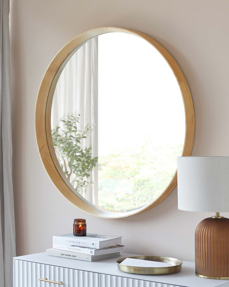 A modern, oval wooden mirror with a natural finish hanging on a wall above a stylish white console table with ribbed detailing. On the table rests a contemporary, ribbed, terracotta-coloured table lamp with a cream shade, alongside stacked books, an amber glass jar, and a golden oval tray. The setup creates a sophisticated and warm aesthetic.