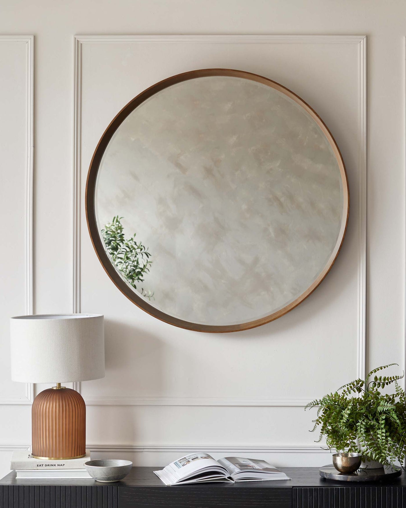 A modern ribbed table lamp with a cylindrical base in a copper finish topped with a white drum shade, placed on a sleek black sideboard with a horizontally ridged design. A potted fern adds a touch of greenery to the setup, accompanied by an open magazine for a casual, lived-in look. The ensemble is set against a neutral wall with classic panelling, complemented by a large round mirror with a subtle, warm-toned frame.