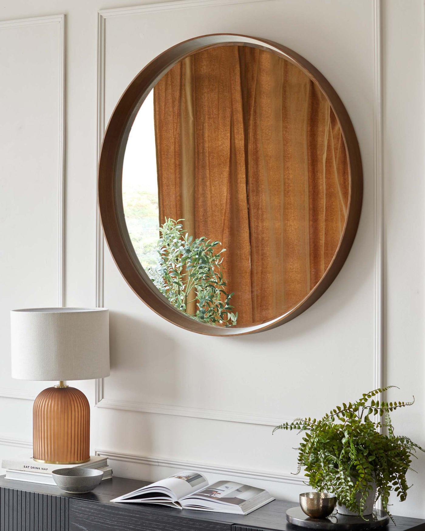 A modern ribbed ceramic table lamp with a cream-colored drum shade, set on a black wooden sideboard with minimalistic styling. A round, wall-mounted decorative mirror with a wooden frame and metallic trim is visible above. A potted fern and an open magazine are also atop the sideboard, adding organic touches to the scene.