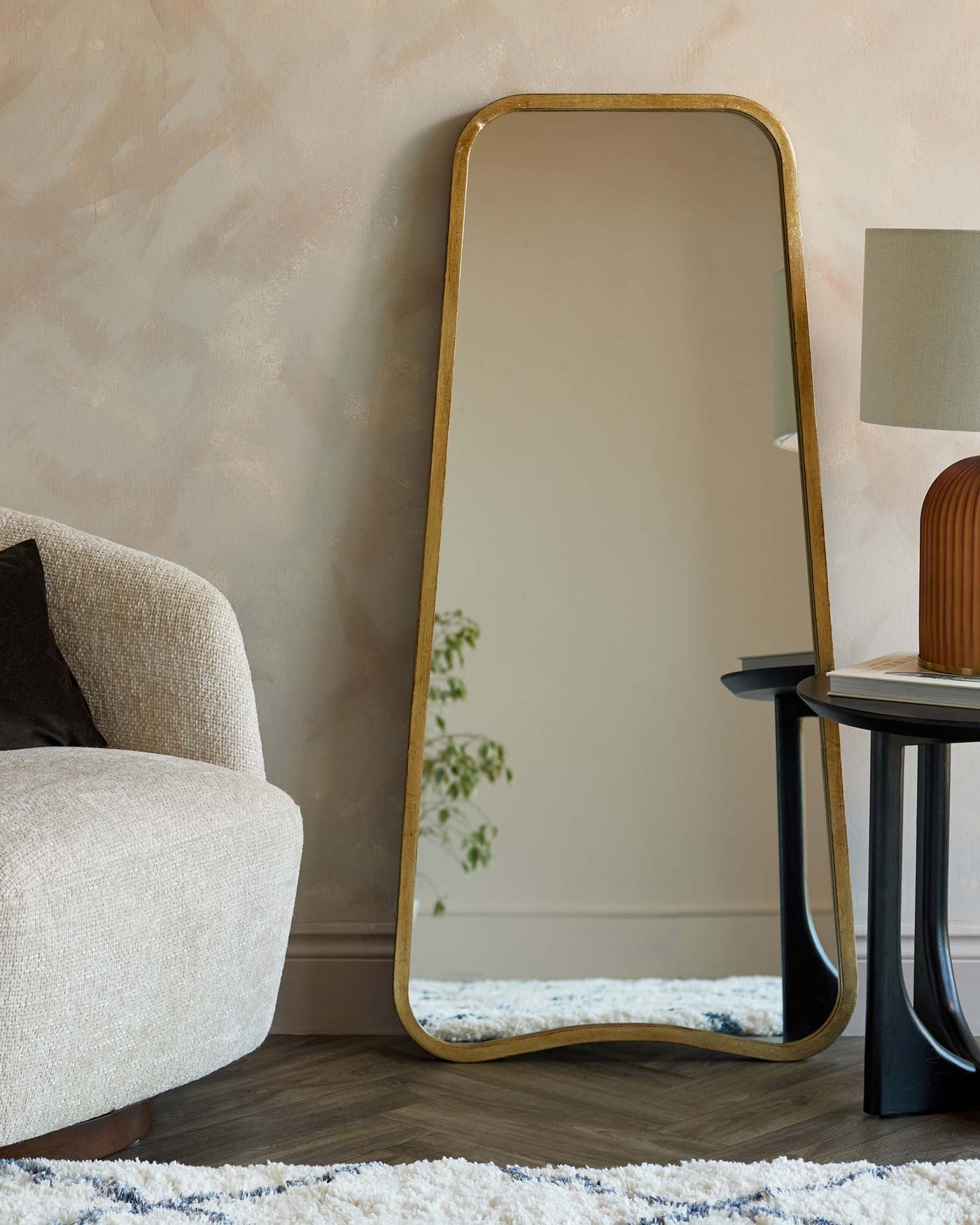 Full-length rectangular floor mirror with a curved top and a thin gold frame, a cream-textured fabric armchair with a dark rounded wooden leg visible, and a round black side table with a cylindrical base and a beige lamp with a white shade. The items are situated on a textured blue and white area rug with a herringbone wood floor visible.