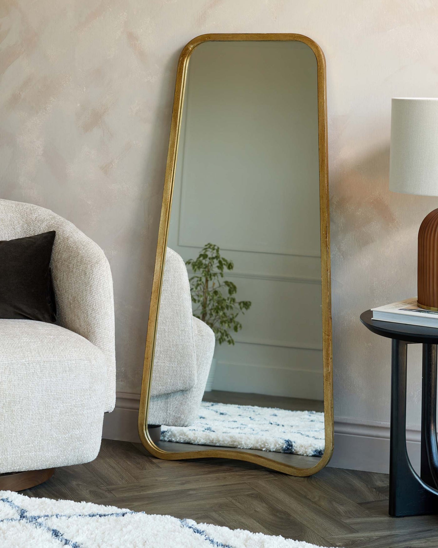 A modern, plush, off-white textured armchair with a black pillow to the left, a large free-standing arched mirror with a slim golden frame leaning against a textured wall, an elegant black side table with a circular top and three slender, slightly splayed legs to the right, and a textured white and blue rug on the floor.
