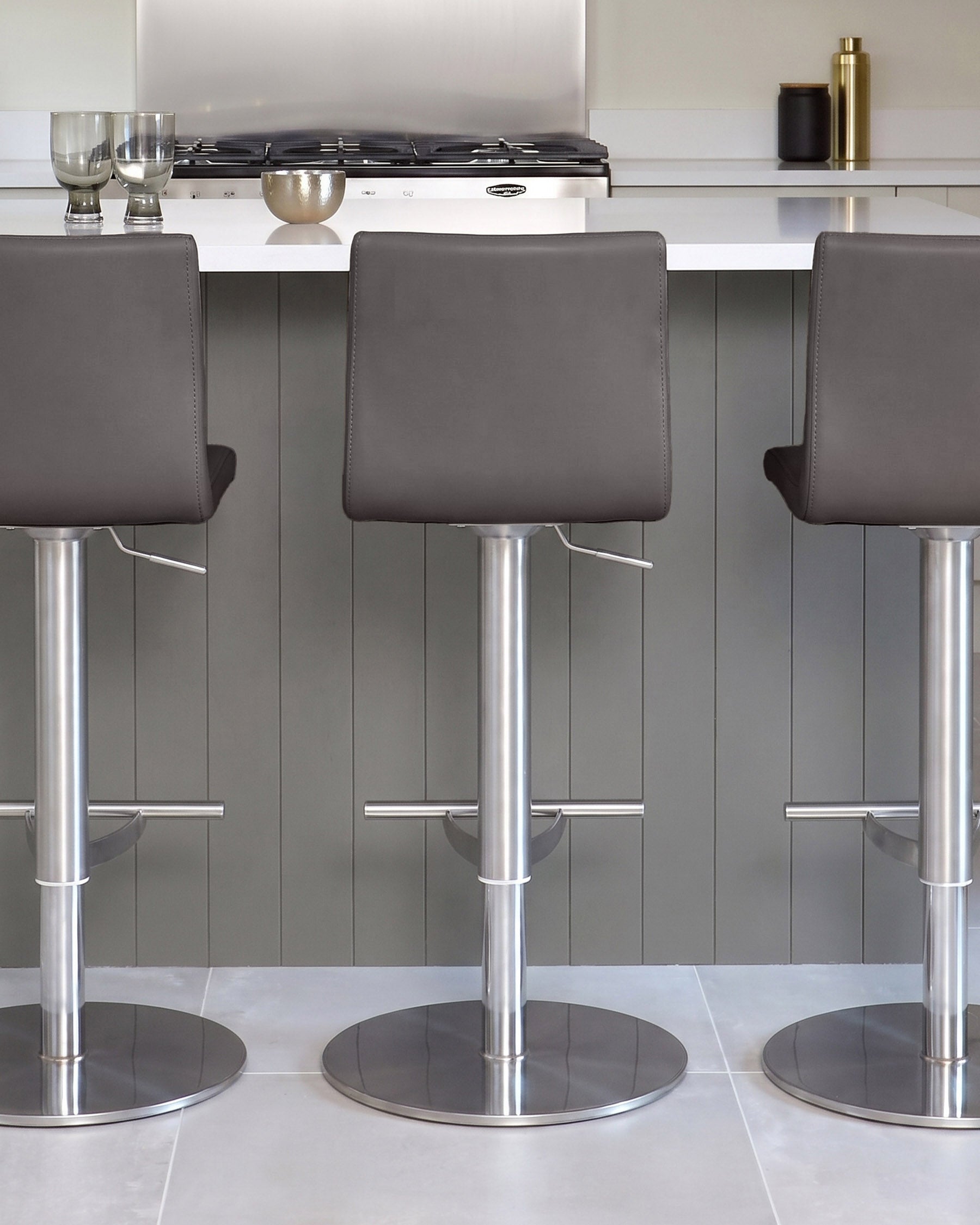 Kiki dark grey real leather gas-lift bar stools with polished stainless steel cylindrical bases, shown in kitchen setting.