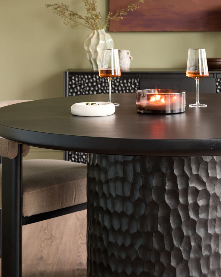 Black round dining table with textured pedestal base, styled with wine glasses, candle, and place settings.