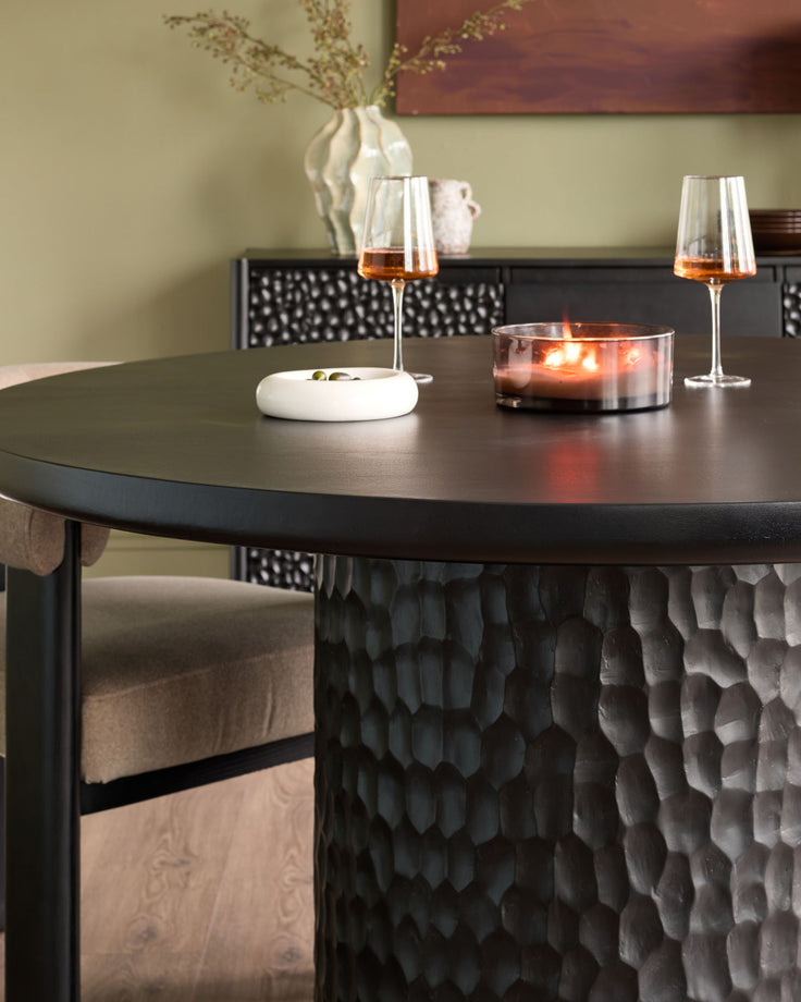 Black round dining table with textured pedestal base, styled with wine glasses, candle, and place settings.