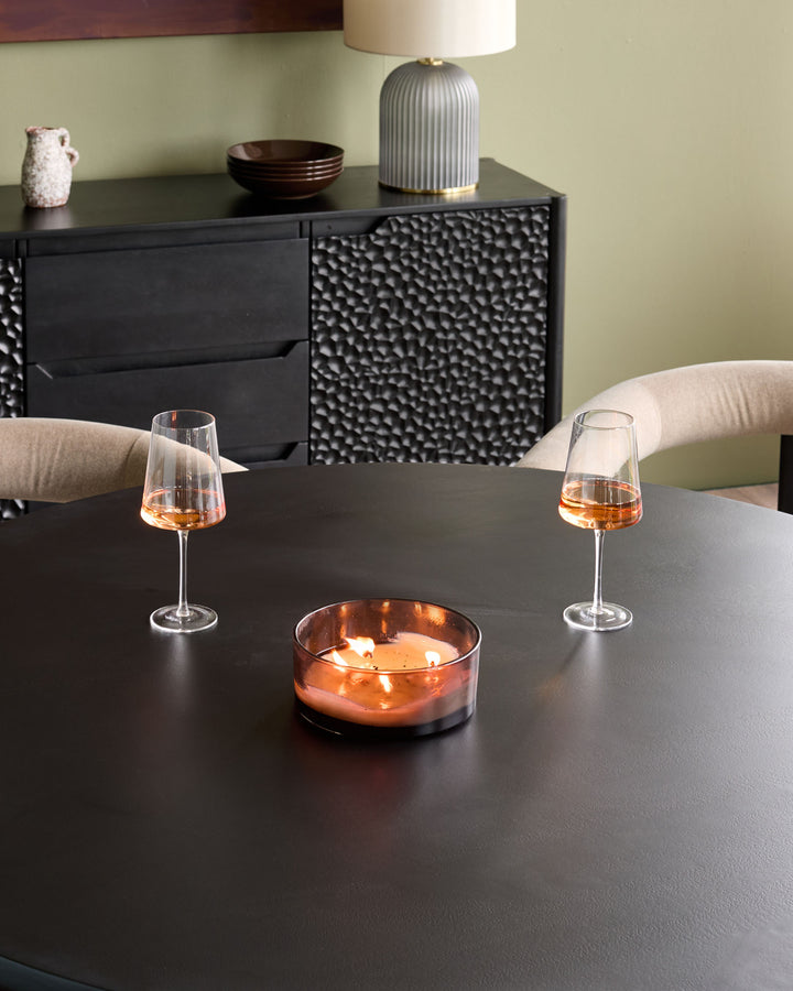 Black wood dining table set for six with two wine glasses and a lit candle, featuring textured cabinet storage behind.