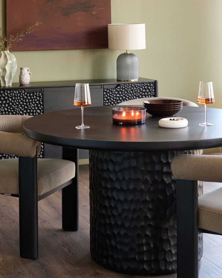 Knox black round dining table for six with textured pedestal base, styled with beige chairs and amber glassware.