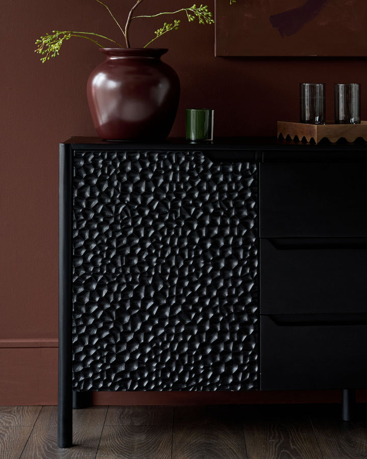 Knox large sideboard in black wood with textured pebble-effect front panel, shown styled with burgundy vase and glassware.