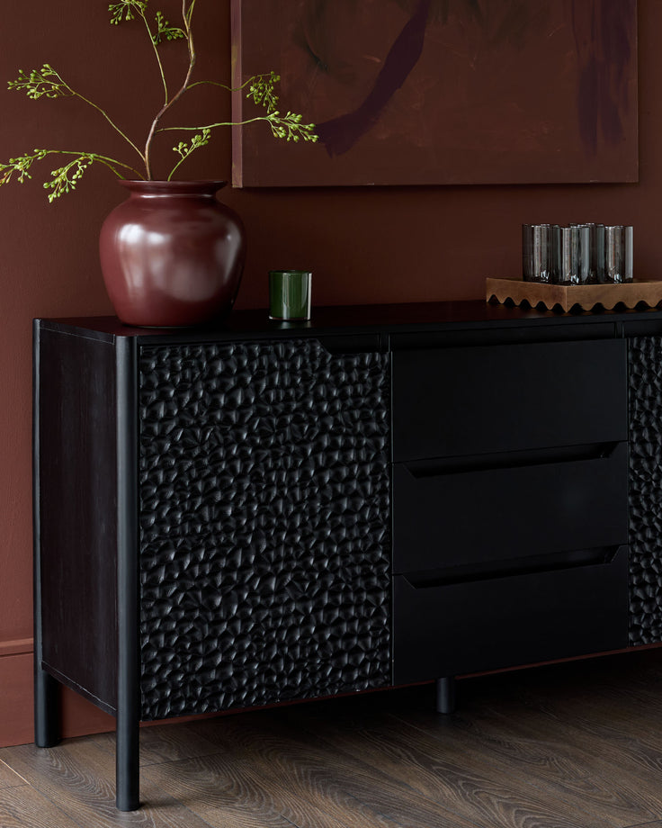 Black wooden sideboard with textured front panels, featuring drawers and doors, displayed against a burgundy wall with decorative vase and greenery.