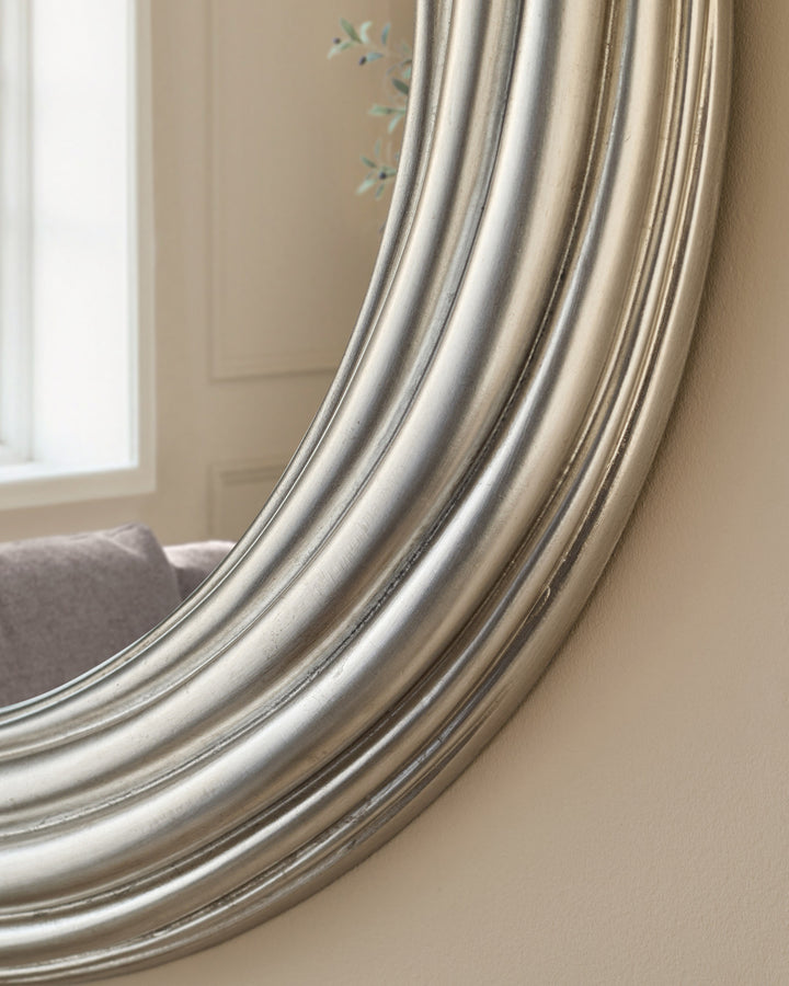 Round silver mirror with layered metallic frame, shown at an angle against a beige wall with window and chair visible.