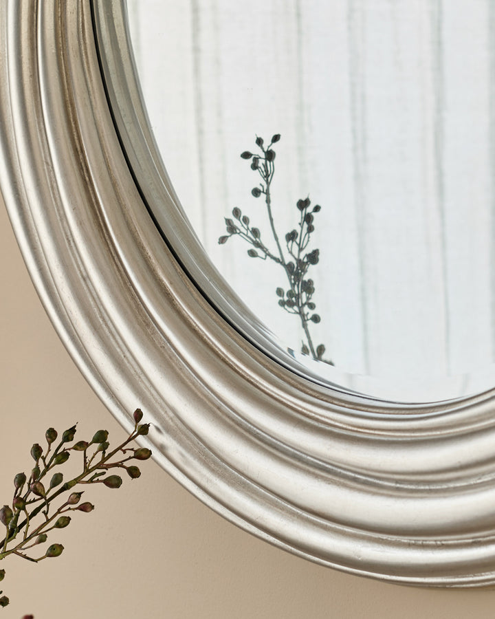 Silver round mirror with decorative rippled frame, displayed against a white wall with dried botanicals.
