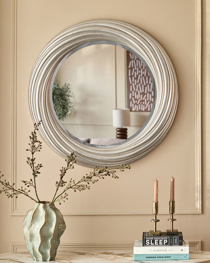 Silver round mirror with concentric ridged frame, hung on beige wall above console table with decorative vase and books.