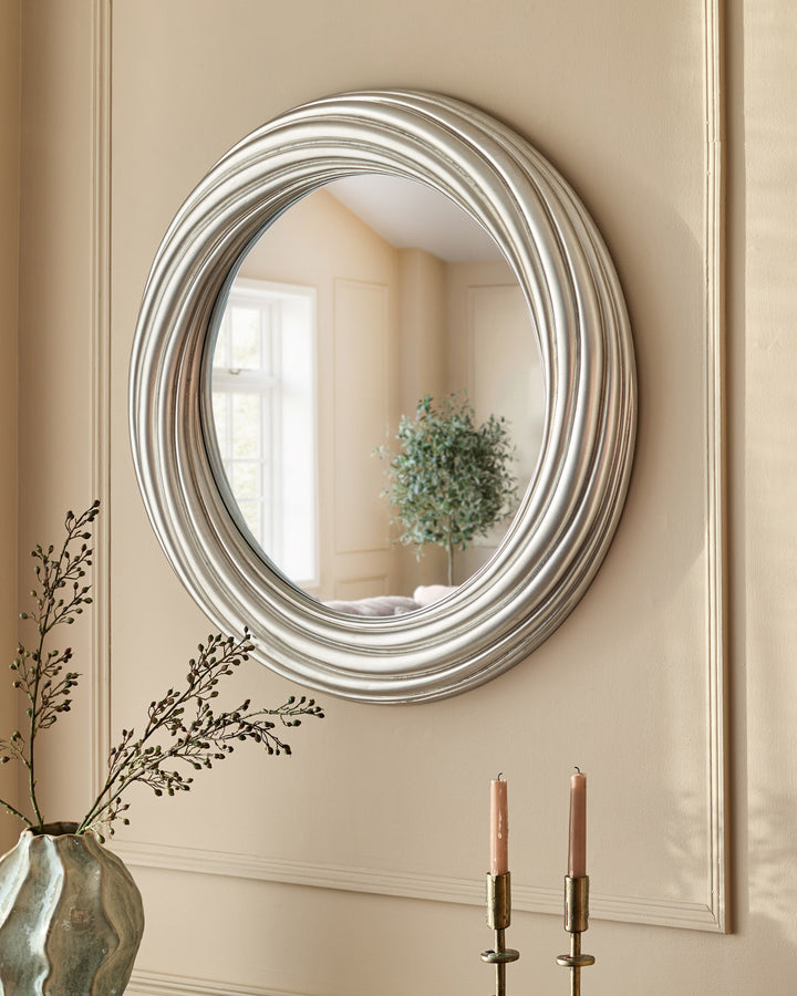 Silver round mirror with concentric circular frame, displayed on neutral wall above candlesticks and decorative vessels.