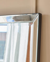 Large leaner mirror with black frame and beveled silver edge trim, angled against beige wall.